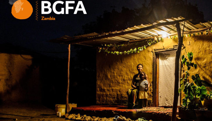 Young girl standing next to her mum outside their house. It is dark outside and a solar powered light bulb is lighting up their veranda.